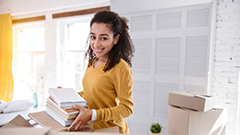 College materials. Charming curly-haired girl holding a pile of books and posing for the camera while packing for moving out of the dorm