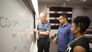 The instructor is writing on a whiteboard with a black marker, and two students are standing next to that person.