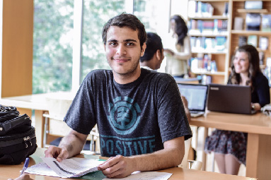 One student is sitting in the library and smiling. He is holding a book. There are a bunch of students in the background using laptops.