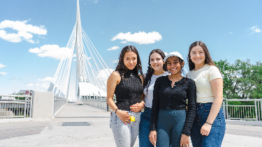 Four students standing in-front of the famous Winnipeg bridge, smiling and looking at the camera.