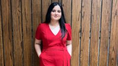 Image of Kamal in a red dress standing in front of a wooden wall